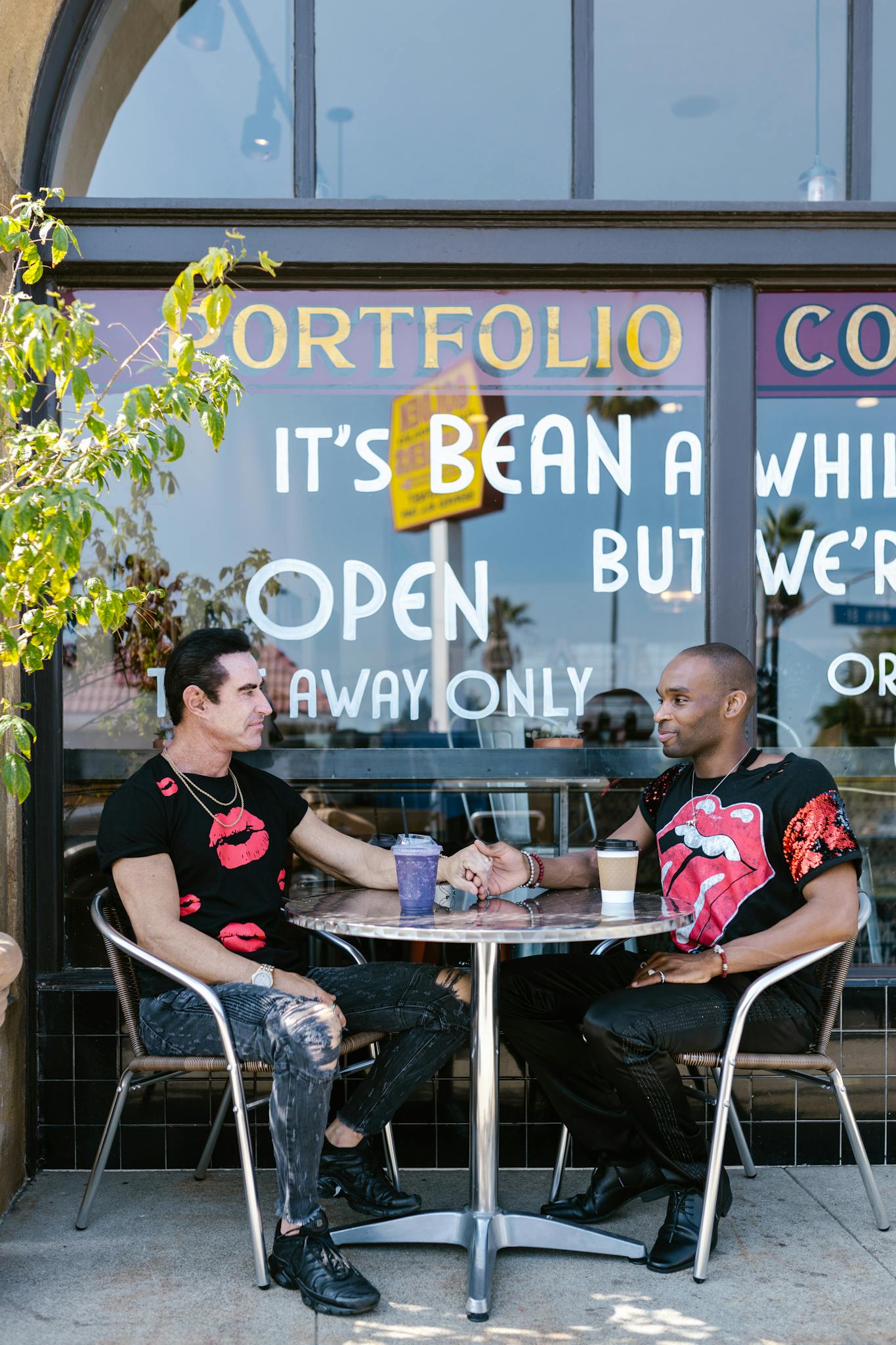 An affectionate interracial gay couple sitting outdoors at a café, holding hands and enjoying their time together.
