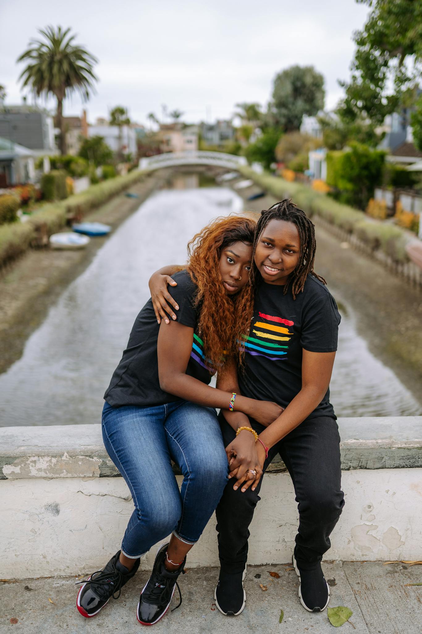An affectionate lesbian couple sits together by a tranquil canal on a summer day, expressing love and unity.